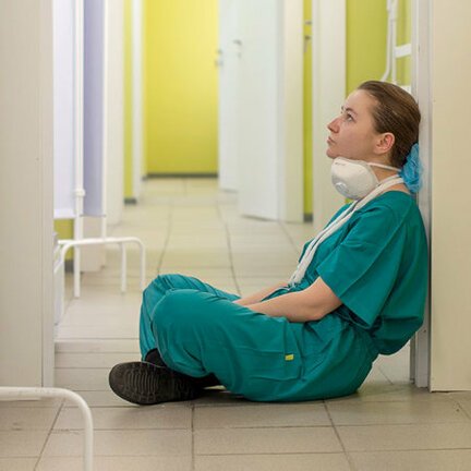 Tired COVID-19 nurse sitting in hallway with mask down and teal scrubs on